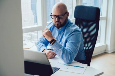 Resmi kıyafet ve gözlüklü dalgın kel erkek işçi bilgisayar masasında oturmuş, Netbook 'u tarıyor ve modern ofisteki kripto para piyasasını inceliyor.