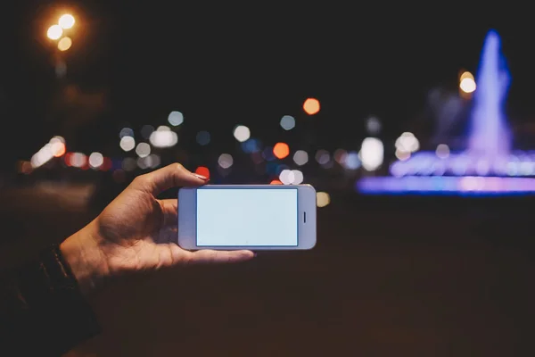 Crop unrecognizable person in black jacket using smartphone with white blank screen and taking photo of city lights in evening