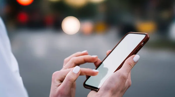 Side view of anonymous female blogger in stylish outfit browsing social media on smartphone while spending time on street in evening