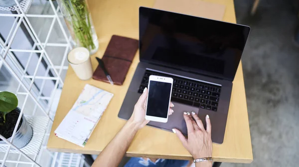 現代のカフェの木のテーブルに座ってコンピュータを操作しながら携帯電話を閲覧するカジュアルな服装で顔のない女性のフリーランスのオーバーヘッド — ストック写真