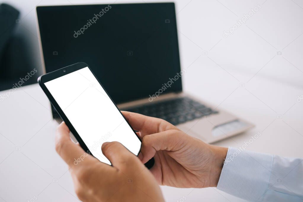 Crop faceless female employee sitting at wooden table with laptop and browsing smartphone with empty screen while working remotely at home