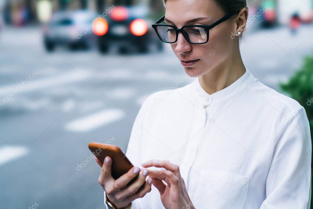 Crop serious young lady in smart casual wear and glasses looking at screen while messaging on digital mobile phone on city street