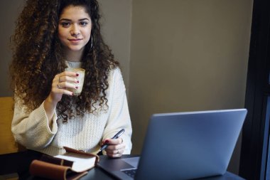 Günlük kıyafetlerle gezen, gülümseyen, kameraya bakan, not tutan ve iş sırasında laptopta kahve içen pozitif kadın serbest yazar.