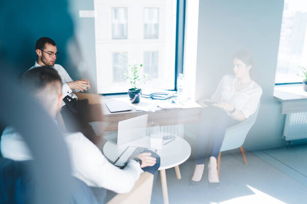 Through window high angle of diverse confident coworkers in formal clothes gathering at table and discussing business project in office