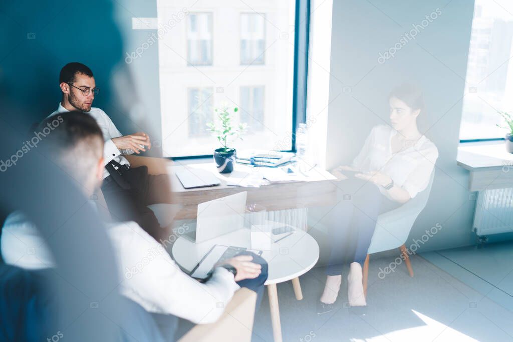 Through window high angle of diverse confident coworkers in formal clothes gathering at table and discussing business project in office