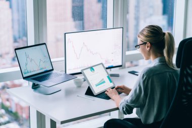 Resmi kıyafet ve gözlüklü odaklanmış kadın girişimcinin ofis içinde kripto para ticareti yapan bitcoin ile çalışmasının yüksek açılı arka görüntüsü