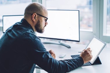 Tablet üzerinde notlar alırken pencerenin yanında monitörlerle oturan şık tişörtlü, odaklanmış erkek çalışanın yan görünümü