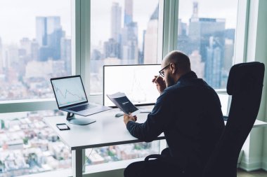 Modern ofiste panoramik manzaralı kağıtları incelerken monitörlerle masasında oturan düşünceli erkek liderin arka planı.