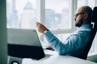 Resmi kıyafetler ve gözlük takan, masa başında oturup, geniş bir ofiste çalışırken dokümanları okuyan konsantre erkek işçilerin yan görüntüsü.