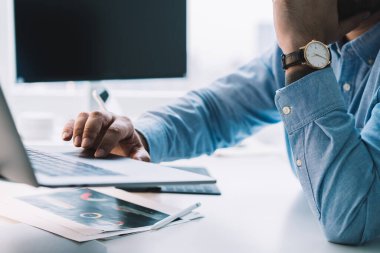 Mavi gömlekli ve kol saati takan ekili adam bilgisayar masasında oturuyor ve ofiste dizüstü bilgisayarla çalışırken dokunmatik ped kullanıyor.