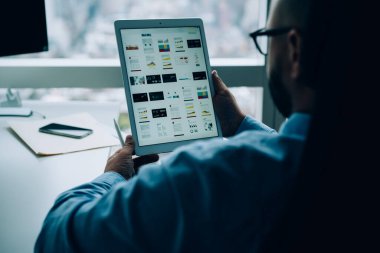 Pencere kenarındaki masada oturan ve karanlık ofiste çalışırken tablet ekranına bakan ekinsiz erkek girişimcinin arka planı