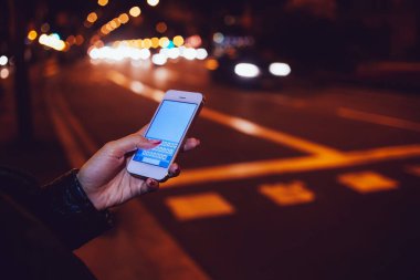 Siyah deri ceketli, meçhul bir kadın gece vakti arabalarla yol kenarında dikilirken modern cep telefonlarına göz atıyor.