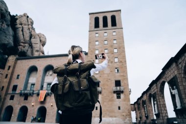 Arka planda sırt çantalı kadın turistin akıllı telefonuyla selfie çekerken Montserrat dağlarını ve Katalonya 'daki manastırı keşfettiğini görüyoruz.