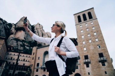 Montserrat Manastırı 'nın ve Katalonya' daki kayalık dağların fotoğrafını çekmek için modern cep telefonu kullanan gündelik giysili sarışın bir kadının alçak açısı.