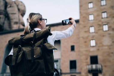 Sırt çantalı kadın turistin Catalonia 'da Avrupa' da seyahat ederken Montserrat manastırını modern cep telefonuyla fotoğraflamasının arka planı.