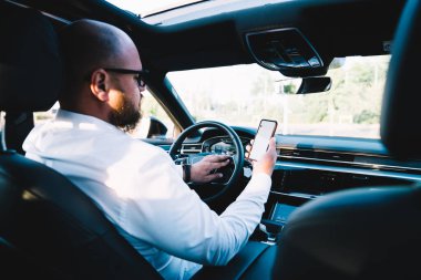 Resmi kıyafetli kimliği belirsiz bir erkek arabanın ön koltuğunda oturuyor ve araba kullanırken güzergahı kontrol ediyor.