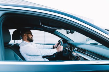 Şoför koltuğunda otururken siyah derili, beyaz gömlekli neşeli erkek yöneticinin yan görüntüsü.