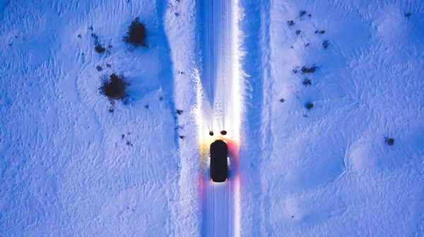 Aerial top view of car on rural area road while headlights are on in winter darkness, bird\'s eye view of suv vehicle in snowy north lands. Couple standing front automobile which lighting them way