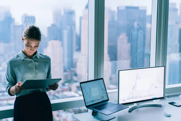 From above of focused female executive manager in formal suit standing near window in office and browsing tablet during work