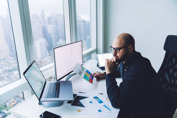 Side view of concentrated bearded male employee with eyeglasses contemplating about financial strategy while sitting at table with gadgets and documents with graphs