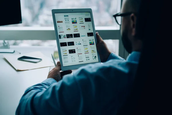 Back view of crop faceless male entrepreneur sitting at desk near window and looking at screen of tablet while working in dark office