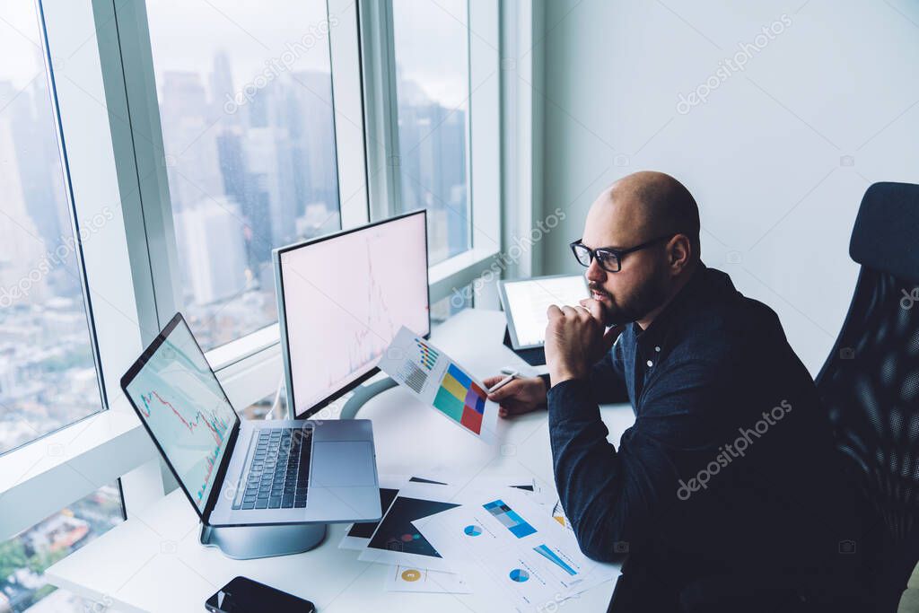 Side view of concentrated bearded male employee with eyeglasses contemplating about financial strategy while sitting at table with gadgets and documents with graphs