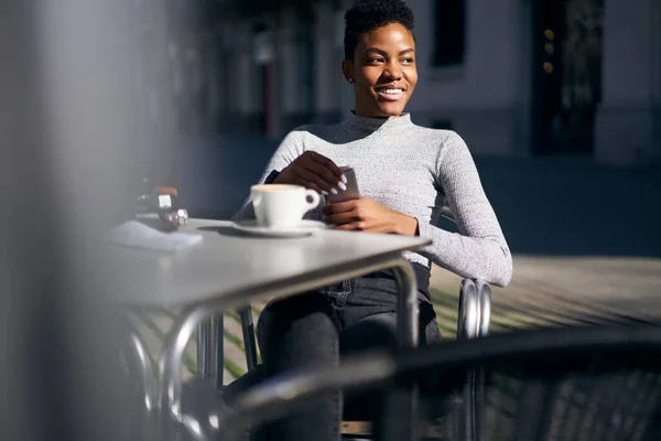 ストリートカフェでコーヒーと写真カメラのカップとテーブルに座っている間に離れて見て スマートフォンを閲覧カジュアルウェアで若いアフリカ系アメリカ人女性を笑顔ミステリー — ストック写真