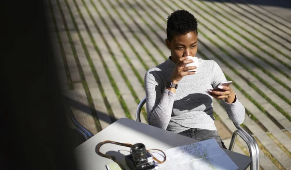 ストリートカフェに座ってコーヒーを楽しみながら スマートフォンの画面に焦点を当てたカジュアルな服の若いアフリカ系アメリカ人女性観光客 — ストック写真