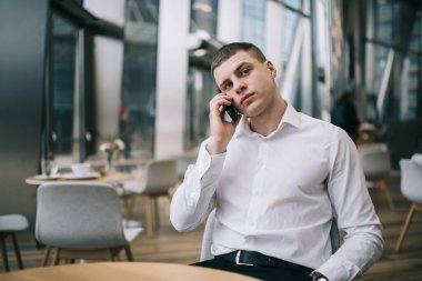 Resmi olarak düşünceli erkek menajer akıllı telefondan konuşuyor ve gündüz vakti modern kafede otururken düşünceli bakışlarını kaçırıyor.