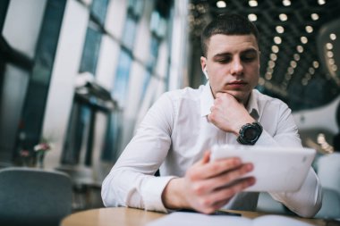 Ofisteki genç yakışıklı adam, modern kafede otururken kablosuz kulaklık takıyor dijital tablette film seyrediyor.