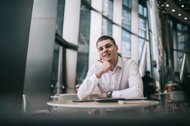 Resmi olarak gülümseyen genç erkek işçi kalem tutarken ve modern kafeteryada günlükle yuvarlak masada otururken ve kameraya bakarken birbirine dokunan bir çene giyer.