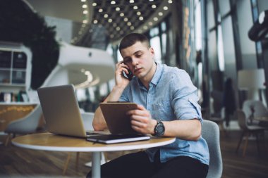 Kafede dizüstü bilgisayarı ve not panosuyla oturup cep telefonuyla konuşan ve proje üzerinde çalışan düşünceli serbest çalışan bir erkek.