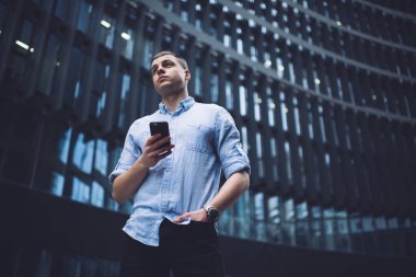 Sıradan giysiler içinde, güncel cam bina önünde el ele dururken akıllı telefon kullanan odaklanmış genç bir erkek çalışandan.