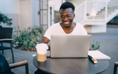 Gündelik giysiler ve gözlüklerle gülümseyen genç siyah bir adam dizüstü bilgisayar kullanıyor ve kahve ile sokak kafesinde oturuyor.