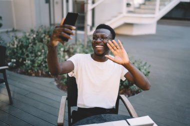 Cheerful dark skinned african american male blogger using smartphone camera for making vlog video sitting on cafe terrace, cheerful african american man waving during mobile phone online call clipart