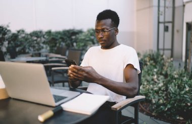 Gündelik giyinen siyahi bir adam, akşam vakti parkta açık bir kafede dizüstü bilgisayarla çalışırken masada oturmuş mesajlaşıyor.