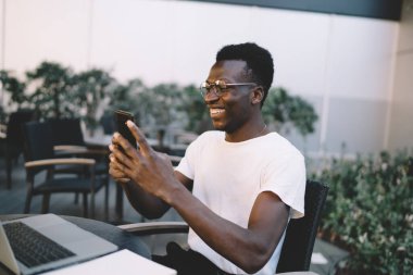 Gülücüklü koyu tenli erkek milenyum göz giysisi haberlerini sosyal ağlardan kafenin terasında akıllı telefonla okuyor. Neşeli Afrikalı Amerikalı adam elinde cep telefonuyla video izlemekten zevk alıyor.