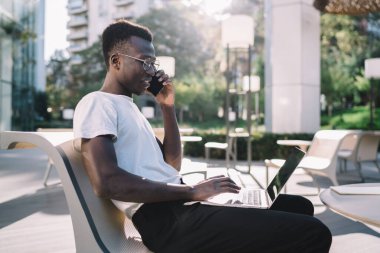 Modern otelin yanındaki terasta dinlenirken akıllı telefonlu ve bilgisayarlı bir sandalyede oturan rahat görünümlü genç siyahi erkek.