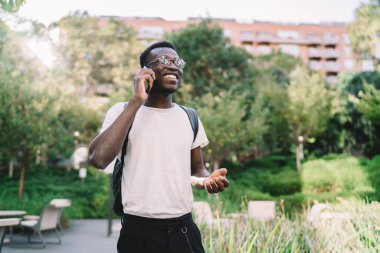 Sırt çantasıyla gezen ve yeşil parkta yürürken telefon görüşmelerine cevap veren ve el kol hareketi yapan tatmin olmuş genç siyahi erkek.