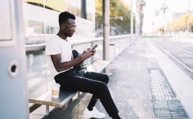 Gündelik giysiler içinde genç koyu tenli bir erkek gezgin akıllı telefondan toplu taşıma tarifesini kontrol ediyor, Afro-Amerikan bir adam cep telefonunda tramvayı bekliyor.