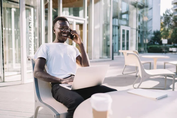 Happy emotional hipster guy freelancer laughing during smartphone conversation working remotely on cafe terrace, smiling prosperous dark skinned male making mobile phone call via roaming