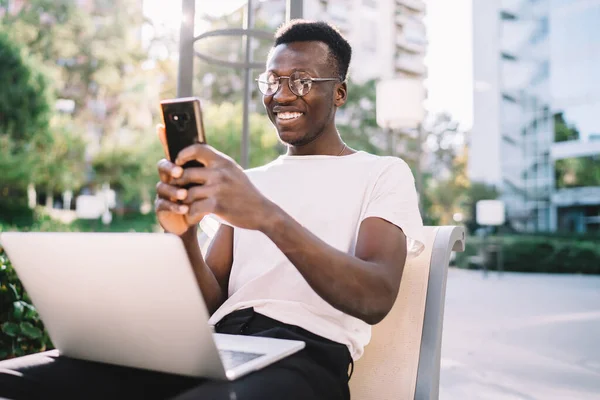 Šťastný Mladý Černoch Brýlích Notebookem Kolenou Zprávy Smartphone Usmívá Zatímco — Stock fotografie