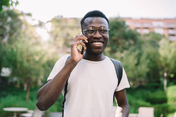 Smiling dark skinned male millennial standing on urban setting talking on mobile phone in roaming, happy african american hipster guy enjoying tariffs for making call in roaming on free time in city