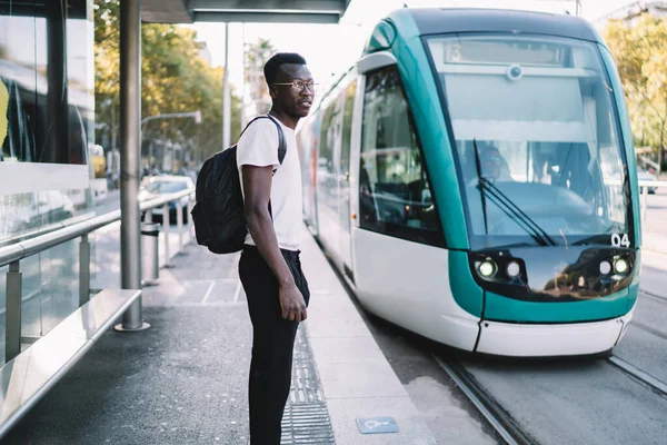 Serious dark skinned hipster guy in casual trendy wear standing on tram station transporting in city, handsome young african american male 20s waiting for modern fast public transport on stop with shelter