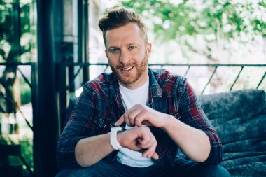 Kendine güvenen kızıl saçlı, günlük giysiler içinde, kafenin açık terasında oturan ve kameraya bakarken bilek saatini gösteren erkek girişimci.