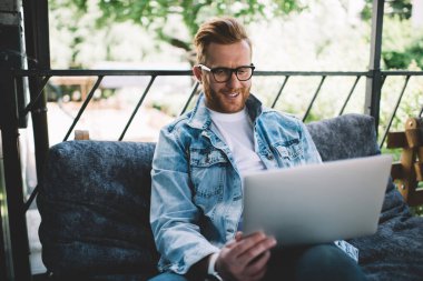 Gözlüklü, yakışıklı, genç ve sakallı bir adam arkadaşıyla konuşuyor. Yaz kafesinde bilgisayarla konuşuyor. Güneş ışığı altında.