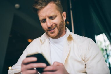 Aşağıdan gülümseyen, kızıl sakallı ve cep telefonuyla mesajlaşan, kafeteryada günlük giysiler giyen bir adamdan.