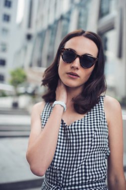 Karizmatik, kendine güvenen, güneş gözlüklü, siyah beyaz kareli elbiseli rahat bir kadın. Bulanık çağdaş binaların karşısında tek başına boynuna dokunuyor.