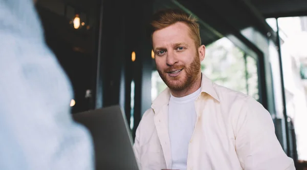 Young cheerful ginger bearded man sitting at table outside cafe smiling, working using laptop during lunch break looking at camera