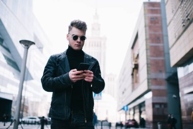 Siyah giysiler ve yuvarlak gözlükler takan düşünceli genç adam sosyal ağda mesajları kontrol ederken sokakta dikilip gökdelenleri ve ofis binalarını sarmış.